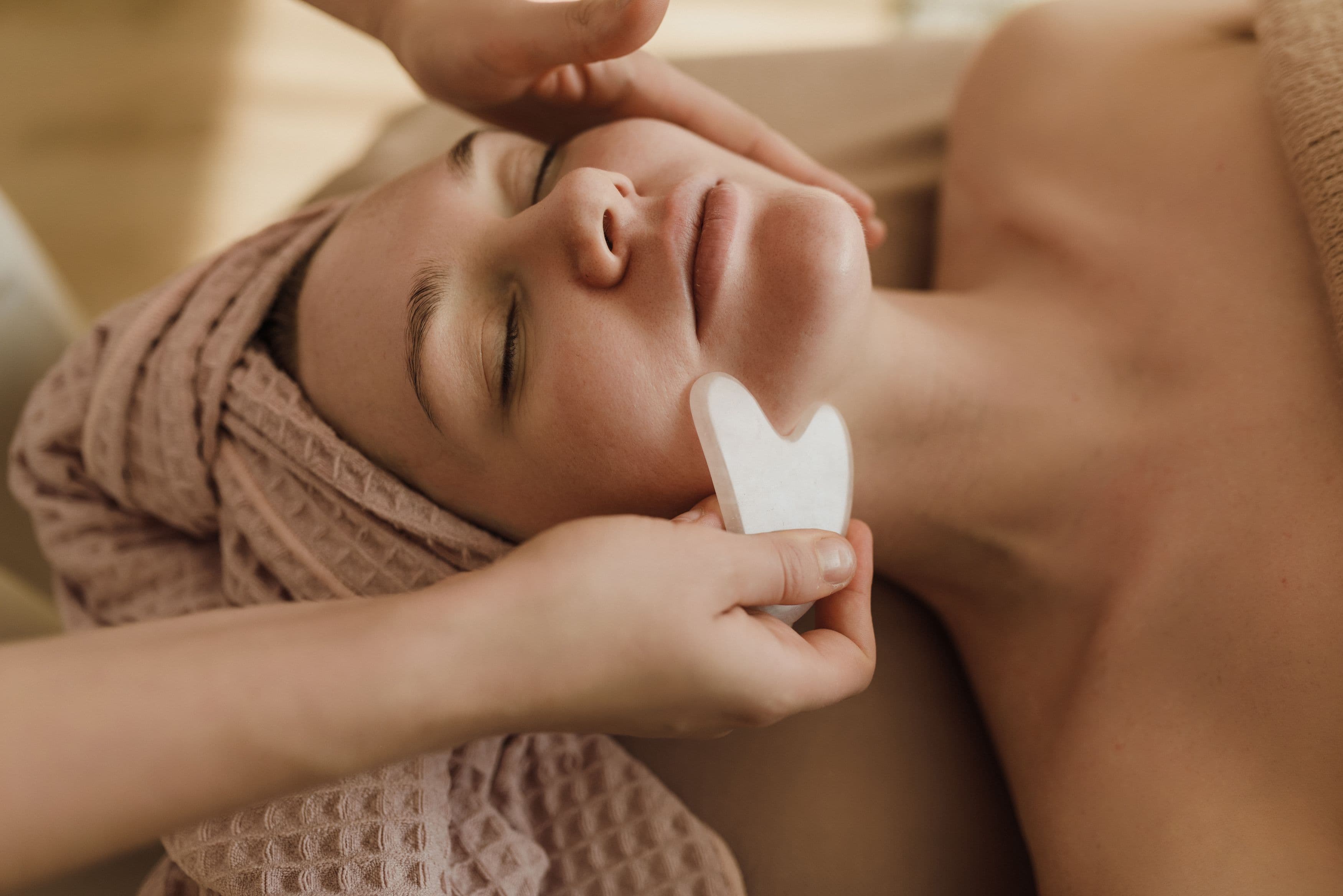 A pale stone gua sha being glided along the cheek during a facial massage.
